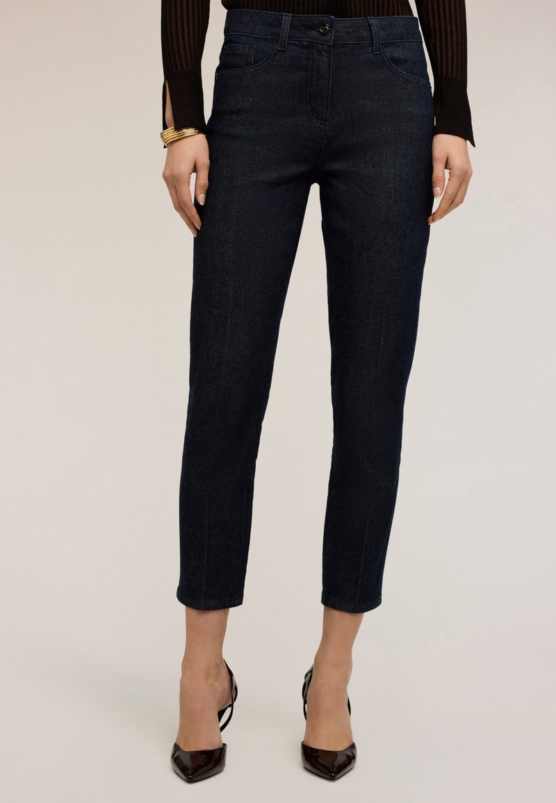 Frau trägt dunkelblaue, hochgekürzte Skinny-Jeans, schwarze spitze Absatzschuhe, ein braunes, geripptes Langarmshirt und goldene Armbänder an einem Handgelenk.