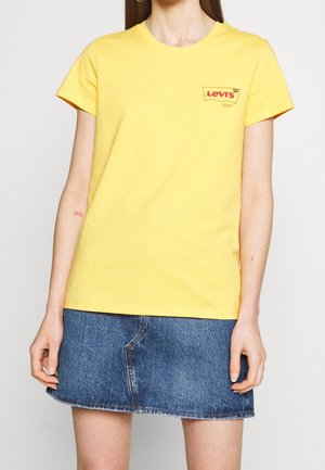 Kvinde iført en gul Levi's t-shirt og et blåt denimsnørk, stående foran en ensfarvet lys baggrund.