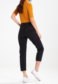 Jeans negros de cintura alta con un ajuste recortado, combinados con una blusa acanalada mostaza. Zapatillas blancas completan el conjunto. Diseño simple y moderno.