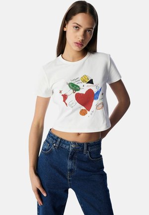 Biała krótka koszulka z kolorowymi doodlowymi ilustracjami, w tym dużym czerwonym sercem i różnymi elementami tekstowymi, zestawiona z niebieskimi dżinsami.