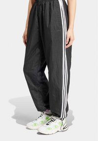 Svarta träningsbyxor med texturerad yta, med tre vita randiga detaljer längs varje ben och en elastisk midja. Matchas med vita och gröna sneakers.