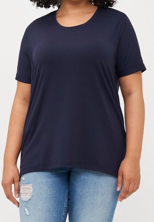 Osoba ubrana w granatowy T-shirt z krótkim rękawem w połączeniu z jasnoniebieskimi podartymi dżinsami, stojąca na jednolitym jasnym tle.