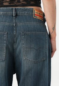 Blugi de denim de culoare albastru închis, cu o textură ușor uzată, având două buzunare pe spate și o etichetă din piele maro cu inscripția "Diesel".