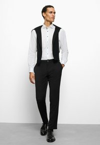 Schwarze, maßgeschneiderte Hose und ein weißes Hemd mit Knöpfen, kombiniert mit einer schwarzen Weste. Verfügt über eine schmale Passform und elegante schwarze Schuhe.