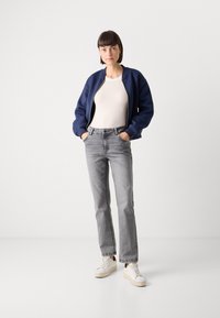 Marine bomberjakke, kremfarget tettsittende topp, lysegrå rettbente jeans og hvite joggesko med beige såler, stående mot en nøytral bakgrunn.