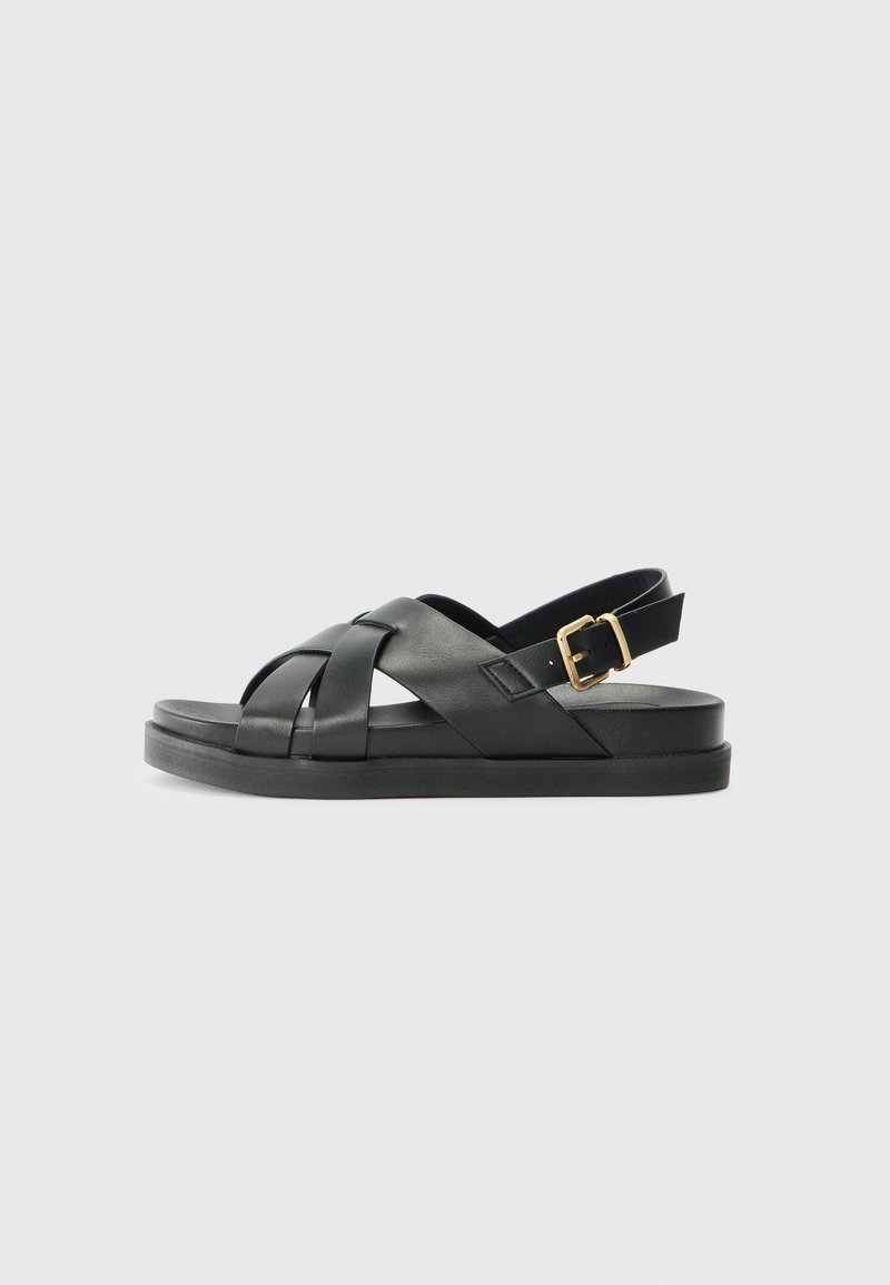 Black leather sandal with a woven strap design, flat sole, and adjustable ankle buckle, featuring a smooth surface and minimalist style.