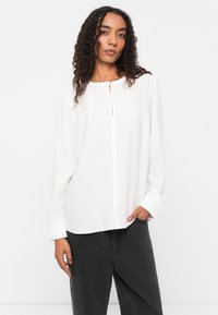 Witte langemouwen shirt met een ronde halslijn en een rits aan de voorkant. De stof heeft een licht gestructureerde afwerking, en de manchetten hebben een gestructureerd ontwerp.