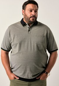Polo grigio a maniche corte con finitura testurizzata, colletto nero con dettagli verdi e taschino sul petto; indossato con pantaloni color khaki.