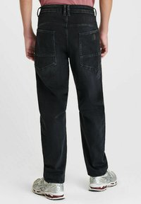 Persoon in zwarte, losvallende jeans met achterzakken en zilveren sportschoenen, staande tegen een effen witte achtergrond.