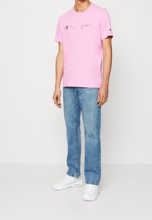 Lyserød bomulds t-shirt med "Champion" logo i et stiliseret skrifttype, parret med lyseblå jeans og hvide sneakers. Sølv kæde-halskæde synligt.