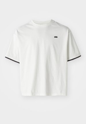Hvit bomull t-skjorte med rund hals, korte ermer og svarte detaljer på ermeendene. Har en liten svart logo på brystet.