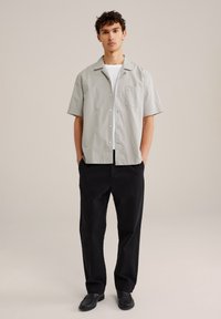 Camisa de botones de manga corta gris claro con bolsillo, combinada con pantalones negros de corte holgado. El modelo lleva unos mocasines negros.