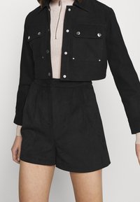 Veste noire courte avec détails de boutons, associée à un short noir taille haute. Tissu texturé avec une finition lisse. Top côtelé rose clair en dessous.