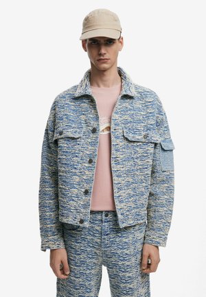 Blue and beige patterned jacket with a collar, button front, and chest pockets, worn over a pink shirt, paired with matching trousers.