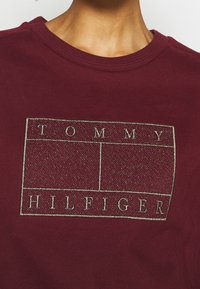 Bordowy sweatshirt z metalicznym złotym logo z napisem "TOMMY HILFIGER" w prostokątnym wzorze. Miękki materiał z okrągłym dekoltem.