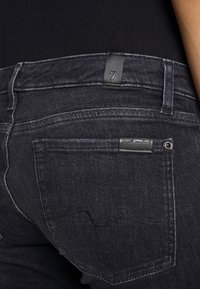 Vaqueros de mezclilla negra con un corte ajustado, costuras sutiles, una etiqueta de cuero con el número '7' y un diseño estándar de bolsillo trasero.