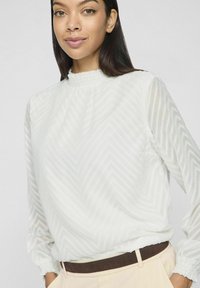 Blusa bianca a maniche lunghe con motivo a chevron texturizzato, scollatura arricciata e polsini elasticizzati. Indossata con pantaloni di colore chiaro.