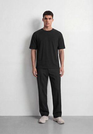 Homme debout contre un mur blanc, portant une chemise noire à manches courtes, un pantalon noir et des baskets de couleur claire, faisant face avec une expression neutre.