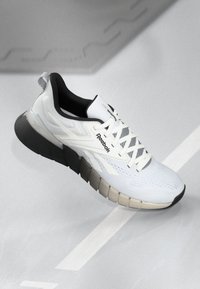 Cască sport albă cu un superior din plasă respirabilă, accente negre, talpă chunky din cauciuc și logo-ul Reebok pe lateral.