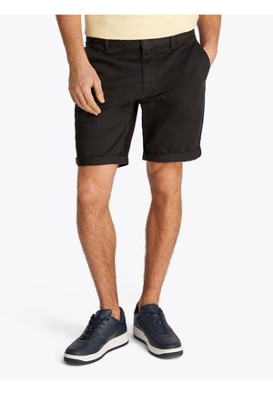 Homme portant un short noir retroussé et des baskets noires, debout avec une main dans la poche sur fond blanc.