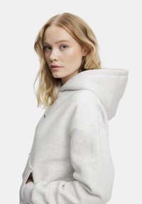 Ljusgrå hoodie gjord av mjukt tyg, med en rymlig huva, kängurufickor och en diskret grön logotyp på bröstet.
