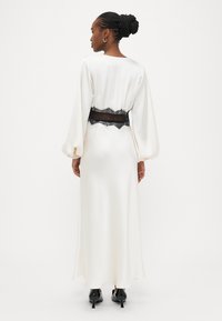 Balta satīna kleita ar garām piedurknēm, melnas mežģīņu jostasvietu. Gluda tekstūra, grīdas garuma dizains un melnas kurpes ar smailiem galiem.