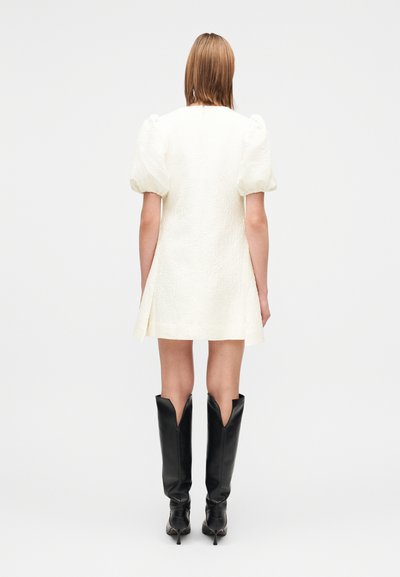 Vestido mini branco texturizado com mangas bufantes, combinado com botas pretas de couro até ao joelho, com recorte em forma de V. Vista traseira.