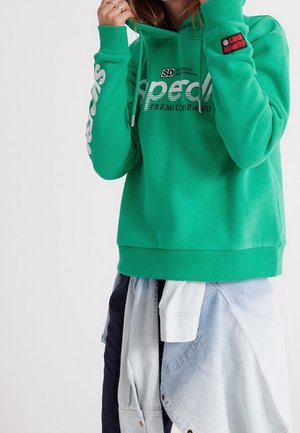 Osoba v jasně zelené mikině s kapucí s bílým textem na hrudi a rukávu, částečně překrývá světle modrou džínovou košili uvázanou kolem pasu.