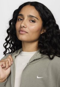 Lys grønn hettejakke med glidelås laget av mykt stoff, med en hvit Nike-logo på brystet. Båret over en lys beige topp med rund hals.