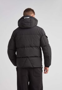 Giacca nera imbottita con cappuccio, caratterizzata da un design trapuntato e polsini elastici. Accentuata da un'etichetta sul retro, abbinata a pantaloni scuri.