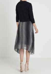 Czarny krótki sweter z rękawami 3/4 zestawiony z midi sukienką w groszki. Sukienka ma przejrzystą warstwę wierzchnią i asymetryczny dół. Beżowe sandały na obcasie.