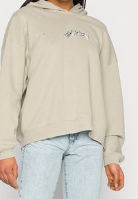 Haut à capuche beige clair avec une coupe décontractée, capuche à cordons et détails brodés argentés. Assorti avec un jean bleu clair, mettant en avant une texture douce.