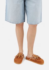 Smeđe suede slip-on cipele s resicama i perlama, u kombinaciji s svijetloplavim trapericama; kratki rub s malo opuštenim krojem.