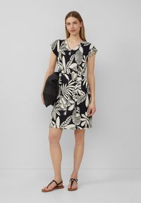 Schwarzes und weißes Blumenmusterkleid mit kurzen Ärmeln, V-Ausschnitt, tailliertem Bund und knielangem Saum. Hält eine strukturierte schwarze Tasche. Sandalen sind zu sehen.