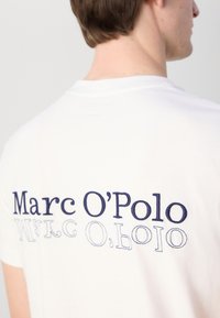 Hvid bomulds t-shirt med mørkeblåt broderet tekst "Marc O'Polo" på ryggen, med varierende skrifttyper og designs.