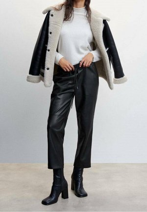 Femme portant un pull blanc, un pantalon noir en similicuir, des bottes à talons noires, et une veste en fourrure noire et blanche drapée sur les épaules.