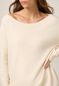 Femme portant un pull ample de couleur crème avec un large décolleté, mettant en valeur ses clavicules, une main posée sur la hanche, cheveux bruns longs flottants.