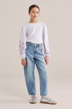 Junges Mädchen trägt ein hellpurpurnes langärmeliges Oberteil mit Blumenstickerei an den Ärmeln, blaue hoch taillierte Jeans und silberne Turnschuhe, steht.