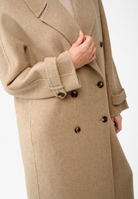 Beige uldblandingsfrakke med dobbeltknap-design, store klapkraver og mørke, runde knapper. Har brede manchetter og en blød struktur.
