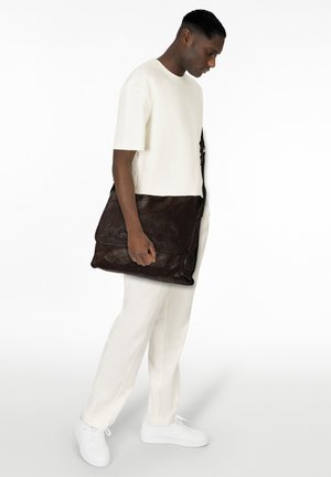 Bruine leren boodschappentas met flapsluiting, gladde textuur en contrasterende stiksels, vastgehouden door een persoon in een witte outfit.