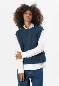 Marineblauer gemusterter ärmelloser Pullover mit Zopfmuster, getragen über einem weißen Hemd mit Kragen, kombiniert mit blauen Jeans.