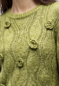 Gros plan d’un pull vert tricoté avec des motifs de feuilles en relief et de petites applications de roses tricotées sur la poitrine et les épaules.