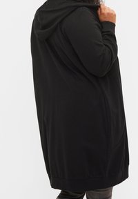 Persona vestida con un abrigo largo negro con capucha y bolsillos laterales, visto desde atrás y sosteniendo la parte de la capucha con una mano.