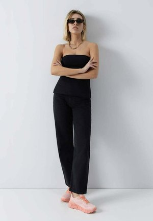 Mujer con top negro sin tirantes, pantalones negros de pierna ancha, zapatillas rosas, gafas de sol y collar de cadena dorada, de pie con los brazos cruzados.