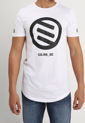 Homme portant un t-shirt blanc à manches courtes avec un grand logo circulaire noir et le texte "CO.RE_XC", associé à un jean bleu.