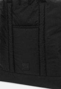 Borsa nera imbottita in nylon con cuciture a filo. Presenta una tasca frontale e un'etichetta con logo testurizzato nella parte inferiore.
