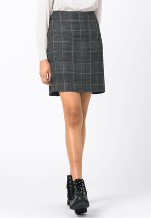 Frau trägt einen knielangen grauen karierten Rock mit einer weißen Bluse und schwarzen Schnür-Stiefeletten vor weißem Hintergrund.