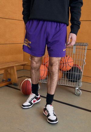 Persoon draagt paarse Los Angeles Lakers basketbalshorts en zwart-witte Nike-sneakers, staat naast een kar met basketbalballen en een houten bank.
