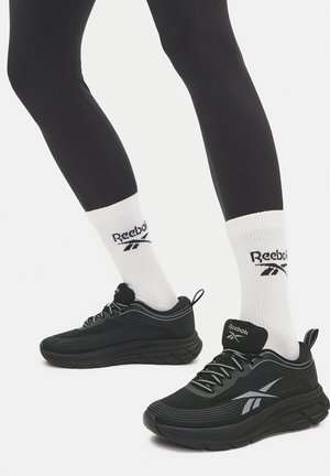 Zapatillas Reebok negras con un diseño texturizado, suela gruesa y detalles reflectantes. Combinadas con calcetines blancos con un logo negro.