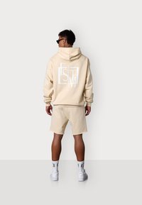 Haut à capuche beige clair avec un logo blanc au dos, associé à un short assorti. Le tissu semble doux, avec un design décontracté et une coupe ample.